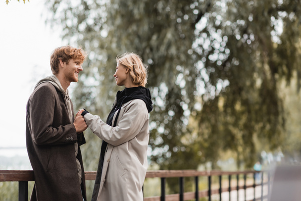 vörös hajú férfi és szőke nő kabátban mosolyog, miközben kéz a kézben a híd közelében a parkban - Fotó, kép