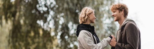 redhead man and blonde woman in coat smiling while holding hands outdoors, banner - Fotoğraf, Görsel