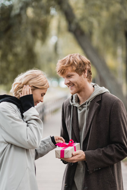 happy redhead man holding wrapped present near pleased girlfriend in park - Photo, Image
