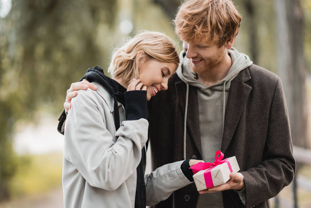 happy redhead man holding wrapped gift box near pleased girlfriend in park - Foto, afbeelding