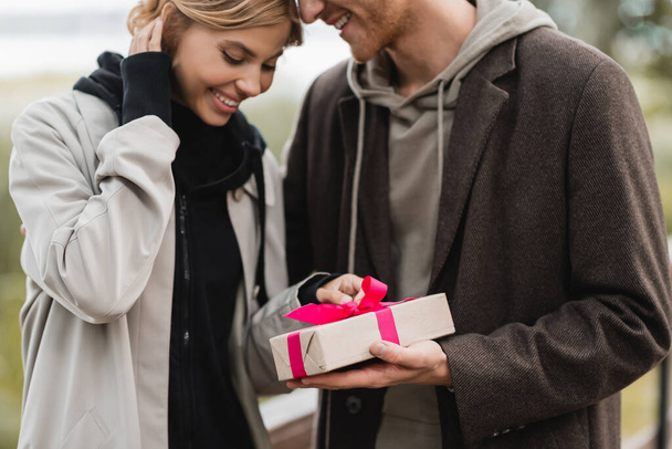 happy man holding wrapped gift box with pink ribbon near pleased girlfriend in park - Фото, изображение