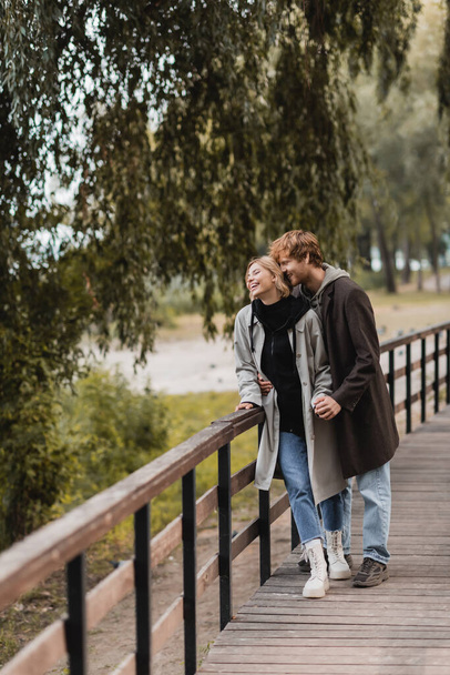 full length of redhead man and blonde woman in coat smiling while having date in park  - Fotó, kép