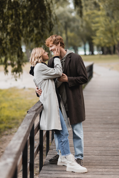 full length of redhead man and blonde woman in coat hugging while having date in park  - Photo, Image