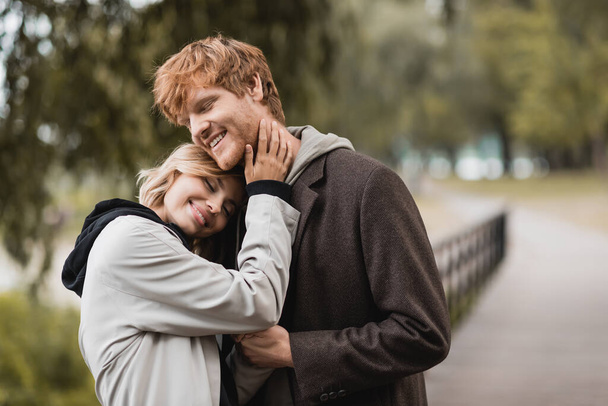 zadowolony ruda mężczyzna i blondynka kobieta w płaszcz uśmiecha się podczas mając datę w parku  - Zdjęcie, obraz