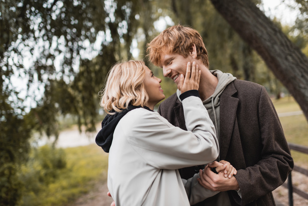 cheerful redhead man and blonde woman in coat smiling during date in park  - Photo, Image