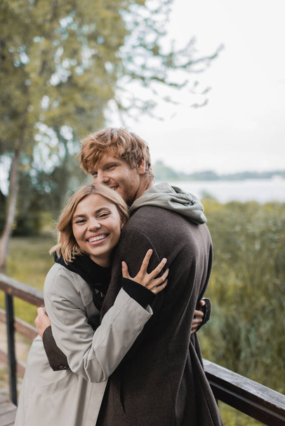 radosna młoda kobieta uśmiecha się i przytula rudego mężczyznę podczas randki na moście w pobliżu stawu - Zdjęcie, obraz