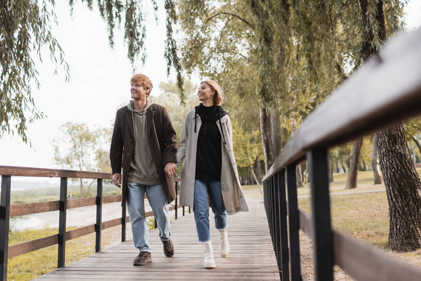 full length of happy couple in coats holding hands and walking on bridge  - Foto, Bild