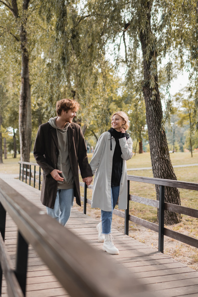 full length of happy couple in coats holding hands and walking on bridge in park - Foto, imagen