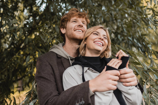 low angle view of happy man hugging cheerful young woman near green leaves on tree - Zdjęcie, obraz