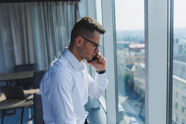 Ένας νεαρός επιχειρηματίας με ένα τηλέφωνο στα χέρια του με γυαλιά και λευκό πουκάμισο, βρίσκεται σε ένα μοντέρνο γραφείο με μεγάλα παράθυρα. Ένας άντρας με κλασικό παντελόνι και λευκό πουκάμισο. Νέος ελκυστικός επιχειρηματίας - Φωτογραφία, εικόνα