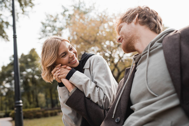 vrolijk en blond vrouw houden hand van roodharige vriend terwijl glimlachen in park - Foto, afbeelding