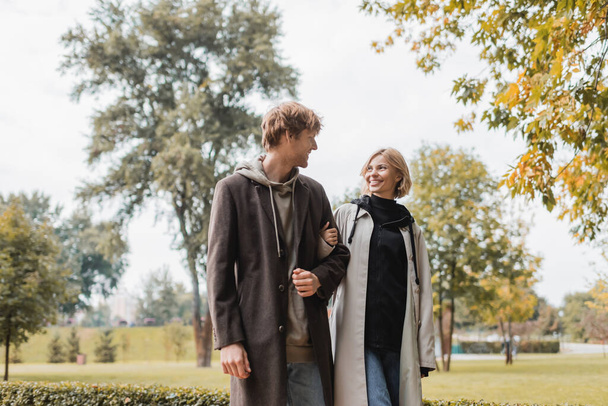 молодая и позитивная пара в пальто, смотрящая друг на друга во время прогулки по осеннему парку во время свидания - Фото, изображение