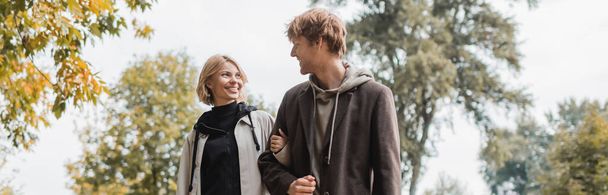 positive couple in coats looking at each other while walking in autumnal park during date, banner - Zdjęcie, obraz