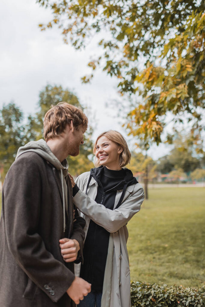 pleased couple in coats looking at each other while walking in autumnal park during date - Photo, Image