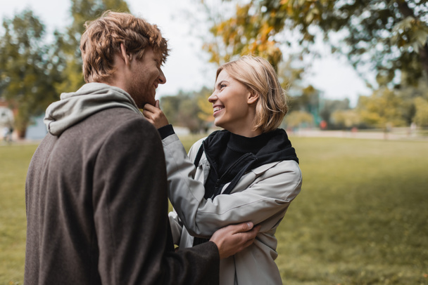 pleased couple in coats looking at each other while hugging in autumnal park during date - Photo, Image