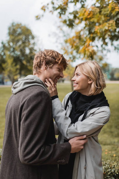 joyful couple in coats looking at each other while embracing in autumnal park during date - Foto, Bild