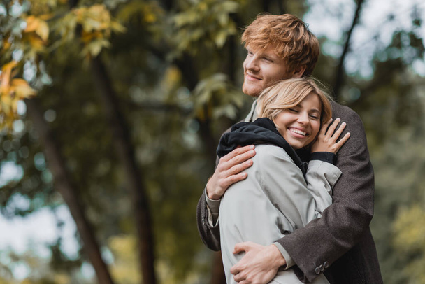 szczęśliwa młoda para w jesiennych płaszczach obejmująca się podczas randki - Zdjęcie, obraz