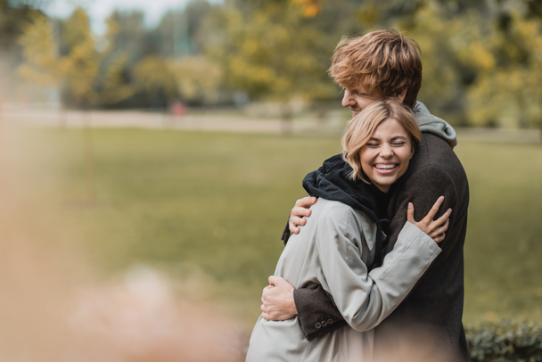 весёлый молодой человек и женщина в осенних пальто обнимают друг друга во время свидания в парке - Фото, изображение