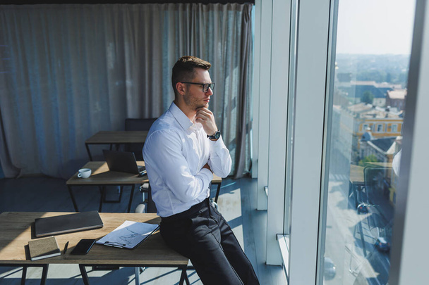 Roztomilý brutální mladý podnikatel v brýlích a bílé košili, je v moderní kanceláři s velkými okny. Muž v klasických kalhotách a bílé košili. Mladý atraktivní podnikatel - Fotografie, Obrázek