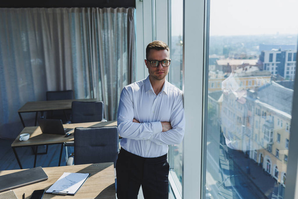 Cute brutal young businessman in glasses and white shirt, he is in modern office with big windows. A man in classic trousers and a white shirt. Young attractive businessman - Фото, зображення