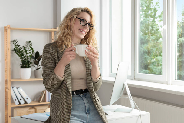 Beautiful young woman with cup of coffee near window in office - Foto, Imagen