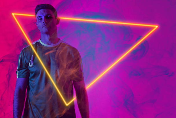 Portrét kavkazského mužského hráče stojícího nad osvětleným trojúhelníkem na neonově růžovém pozadí. Kopírovat prostor, digitální kompozit, sport, fotbal, soutěž, záře, kouř a abstraktní koncept. - Fotografie, Obrázek