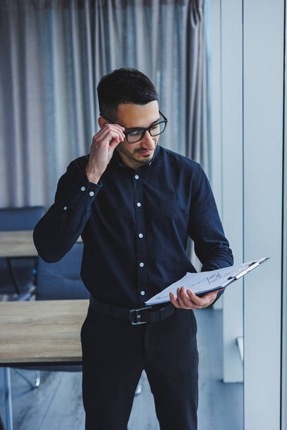 A Caucasian business man in glasses and a black shirt sits in the office and looks at sales growth charts. A young successful businessman in classic clothes stands by the window in the office - 写真・画像