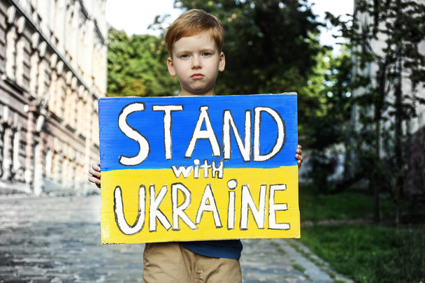 Sad boy holding poster in colors of national flag and words Stand with Ukraine on city street - Foto, Imagem