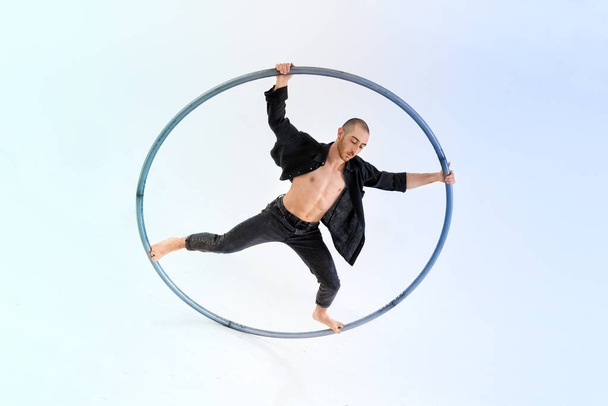 Full body barefoot man in jeans and black shirt doing trick on cyr wheel during performance against gradient blue background - Photo, Image