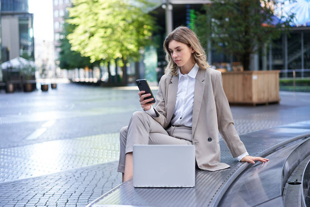 Firemní žena v obleku pracuje v centru města, pomocí notebooku a mobilního telefonu. - Fotografie, Obrázek