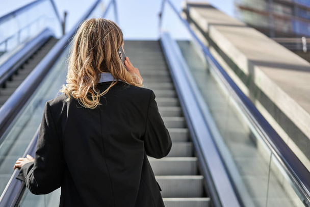 Rear view of businesswoman in suit, standing on escalator and talking on mobile phone. - Photo, image