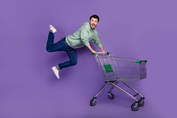 Full body portrait of crazy carefree person jump fly push shopping trolley isolated on purple color background. - Foto, Imagen