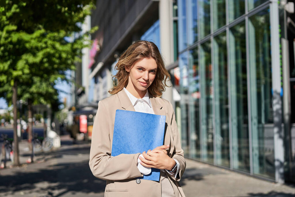 Уверенная улыбающаяся деловая женщина, держащая синюю папку. Студентка в бежевом костюме, держит синюю папку с документами и смотрит в камеру, стоит снаружи офиса. - Фото, изображение