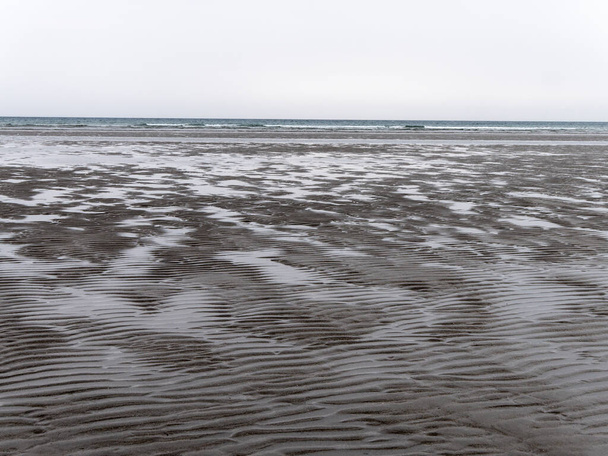 Влажный волнистый песок на берегу моря во время отлива в облачный день. Мрачный морской пейзаж. Жесткая природа севера Европы. Холодное побережье Атлантики. - Фото, изображение