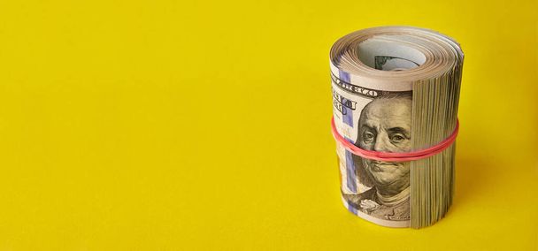 Many hundred dollar bills rolled up with a red rubber band on a yellow background. - Foto, afbeelding