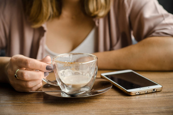 Egy szőke hajú, fehér inges fiatal nő egy csésze tejeskávét vagy cappuccinót tart egy kávézóban. Az okostelefonjával dolgozik. - Fotó, kép