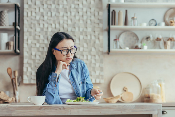 Displeased young Asian woman in glasses and denim shirt sitting in the kitchen at home at the table. He tries a fresh salad with a fork, looks at it, tired of being on a diet. - Foto, imagen