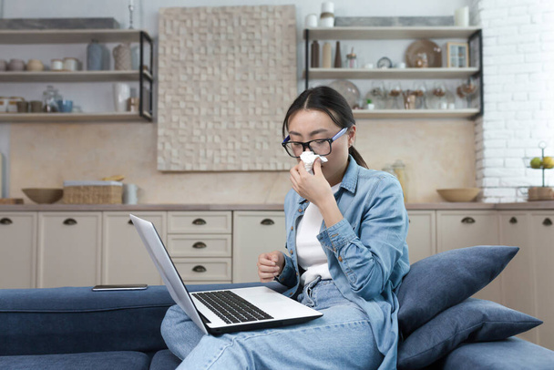 Work from home. Hospital. A young Asian woman freelancer fell ill. Works from home on the sofa at the laptop. He feels bad. He has a runny nose, wipes his nose with a napkin. - Fotoğraf, Görsel