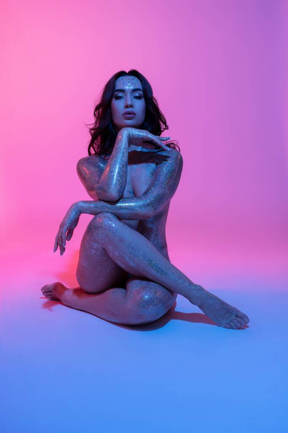 Full body of young sexy lady with glossy paint on body sitting in neon illuminated studio - Foto, Bild