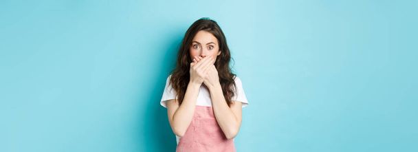 Portrait of shocked glamour girl gasping, covering mouth with hands and staring at camera surprised, hear gossip, standing over blue background. - 写真・画像