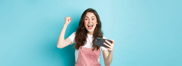 Excited beautful girl winning on mobile phone, holding smartphone and shouting yes with joyful face and fist pump, smiling amazed at camera, blue background. - Photo, Image