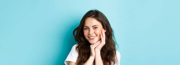 Huidverzorging en schoonheid. close-up van aantrekkelijke jonge vrouw glimlachen met tanden, tonen haar schone gloeiende huid met heldere make-up, staande over blauwe achtergrond. - Foto, afbeelding