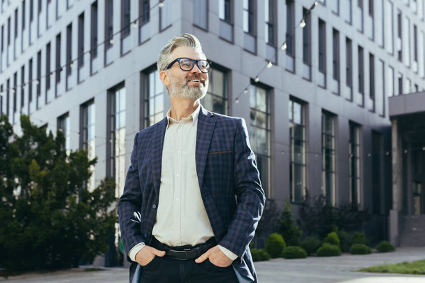 Portrait of senior successful investor, mature businessman outside office building in business suit and glasses, gray haired banker smiling and looking away. - Foto, Imagem