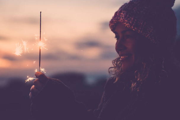 Side view of happy woman using sparkler light and celebrate alone in outdoor leisure activity. Winter season and female people. Christmas or new year eve celebration concept lifestyle. Dark portrait - Photo, Image