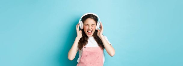 Carefree pretty girl scream from joy, listening music in headphones with happy face, lip-sync lyrics, standing over blue background. - Fotografie, Obrázek