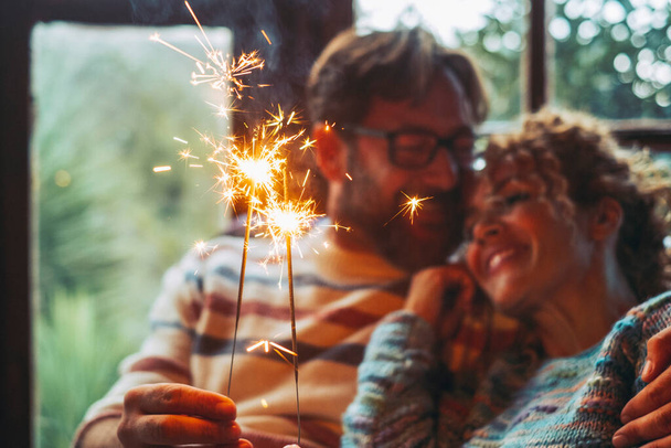 Love and romantic leisure activity with man and woman holding fire sparkler light together hugging and enjoying relationship at home sitting on a sofa. Christmas time for happy couple alone indoor - Photo, image