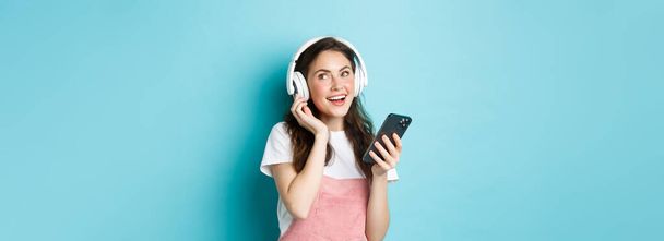 Image of young woman put on headphones and pick music on smartphone, holding phone, listening song, standing over blue background. - Photo, Image