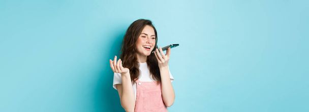 Портрет улыбающейся женщины, держащей телефон у губ и разговаривающей, использующей переводчик приложения на смартфоне или записывающей голосовое сообщение, говорящей на громкой связи, стоящей на синем фоне. - Фото, изображение