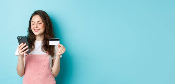 Jeune fille moderne payant en ligne avec carte de crédit, tapez des informations sur le smartphone, regarder l'écran avec le visage heureux, shopping dans l'application, debout sur fond bleu. - Photo, image