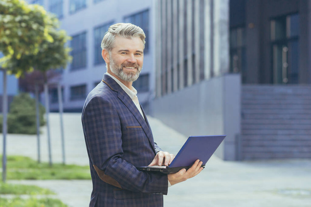 Portrait of mature businessman outside modern office building, senior gray-haired investor with laptop smiling and looking at camera, successful man in business suit holding netbook. - Fotó, kép
